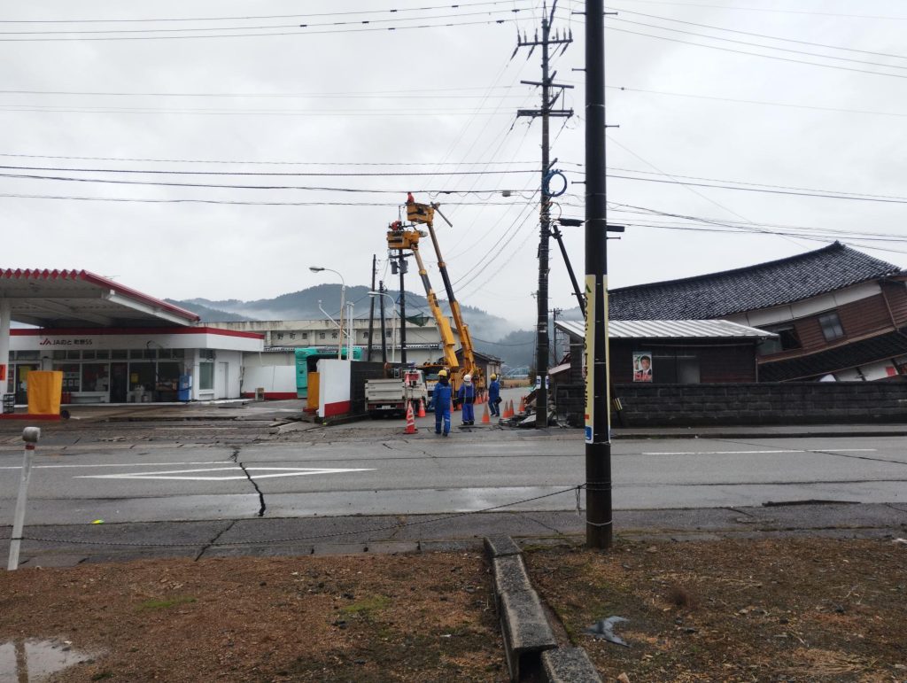 道路には亀裂が入り、家屋は倒壊している。奥に2台の高所作業車がっ作業している。