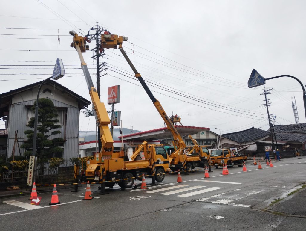 道路上で2台の高所作業車がアームを伸ばして電線を修理。奥には倒壊し家屋が見える。