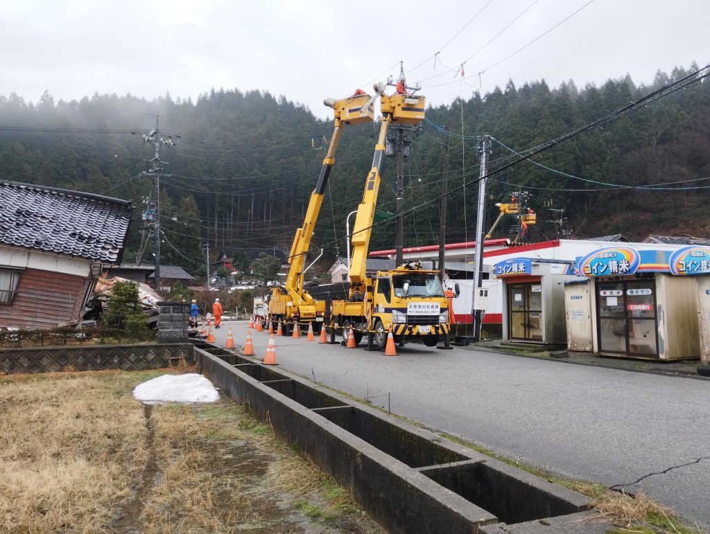 田んぼ脇の道路で2台の作業車がアームを伸ばして電線を修理している。お国も別の2台の高所作業車が作業している。