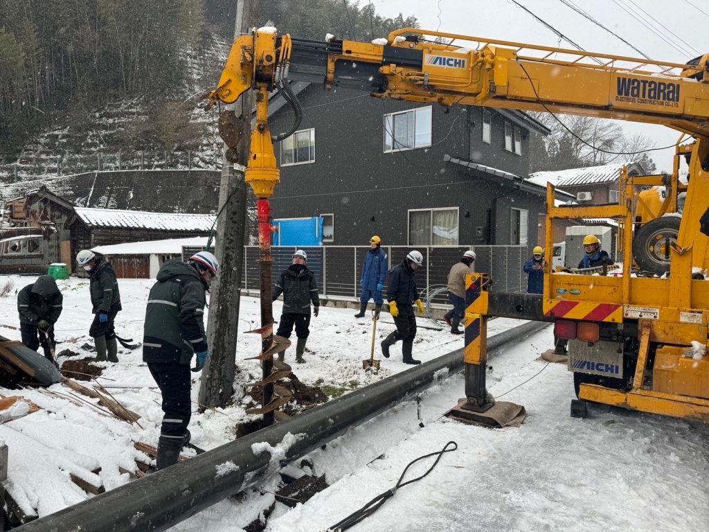 雪が降る中、建柱車が掘削している。作業員数名が作業している。