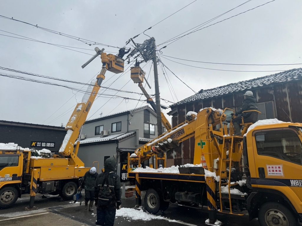 上部が折れている電柱に2台の高所作業車がアームを伸ばしている。手前には建柱車1台が待機している。