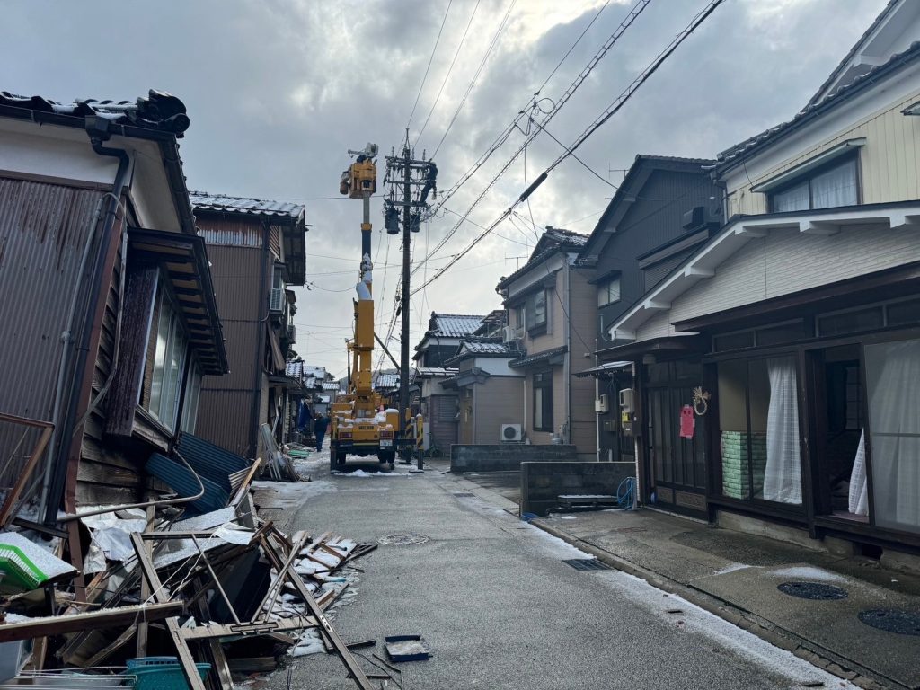 住宅街の電柱横で高所作業車がアームを伸ばして作業している。手前の住宅は1階部分が倒壊している。