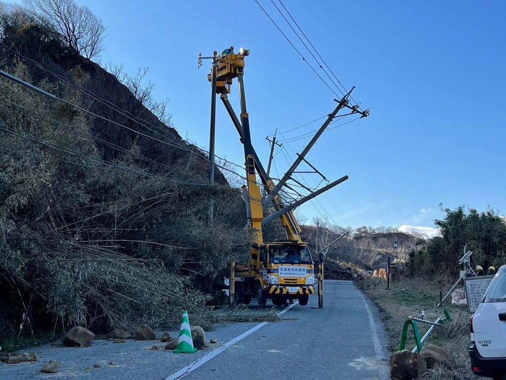 傾いた電柱の横に新しい電柱がまっすぐ立っている。高所作業2台が新しい電柱にアームを伸ばし、作業員が作業している。