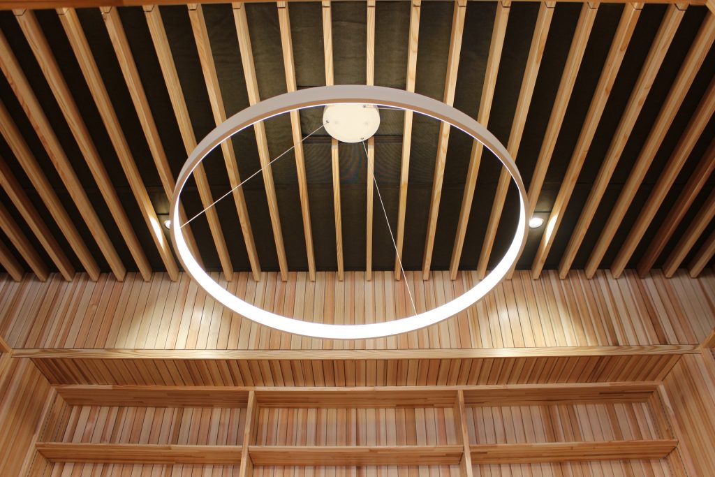 メディアセンターの本棚に囲まれた空間の照明。ルーバーの間にダウンライトがある。天井からは輪の形をしたオブジェが吊り下げられ、その内側が光っている。