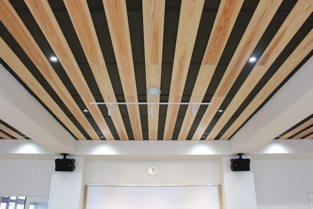 音楽室の天井。黒い下地の上に木製の板が等間隔に設置され、ストライプのような模様。黒い縞の部分にダウンライト、ホワイトボード前にはスポットライトが取り付けられている。