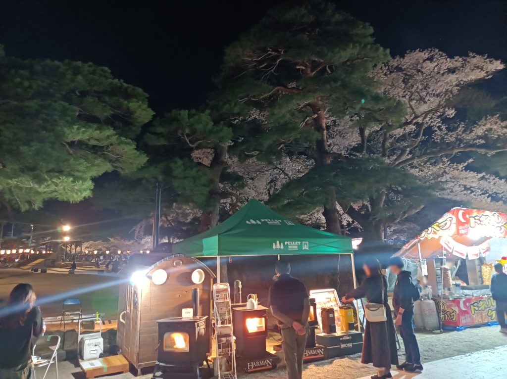 夜、当社展示ブースの風景。4人のお客さんが展示物を見ている。ブースの後ろには松の木と満開の桜の木。