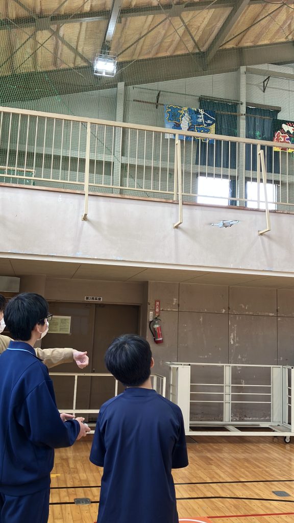 男性社員が操作方法を教えながら男子生徒が操縦し男子生徒がそれを見ている