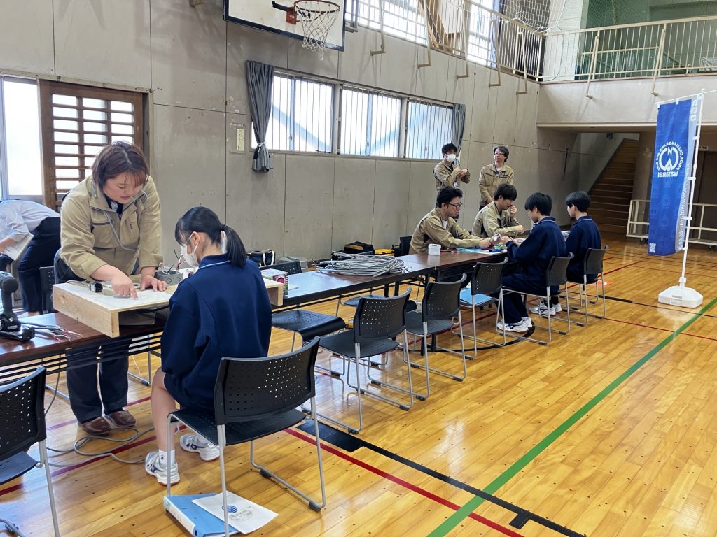 当社の社員3名が生徒3名に説明をしている。後ろに男性社員が見ている